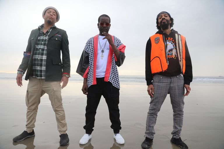 Hip-hop group The Pharcyde three members posing on an ocean beach in full color for an outdoor promotional portrait against an overcast sky.